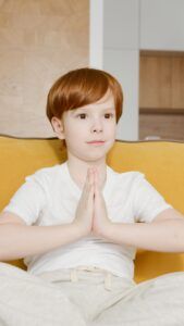 Young caucasian child with red hair in a meditation pose indoors, promoting relaxation and healthy lifestyle.