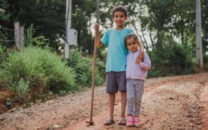 Two children smiling and standing on a rural dirt path surrounded by greenery.